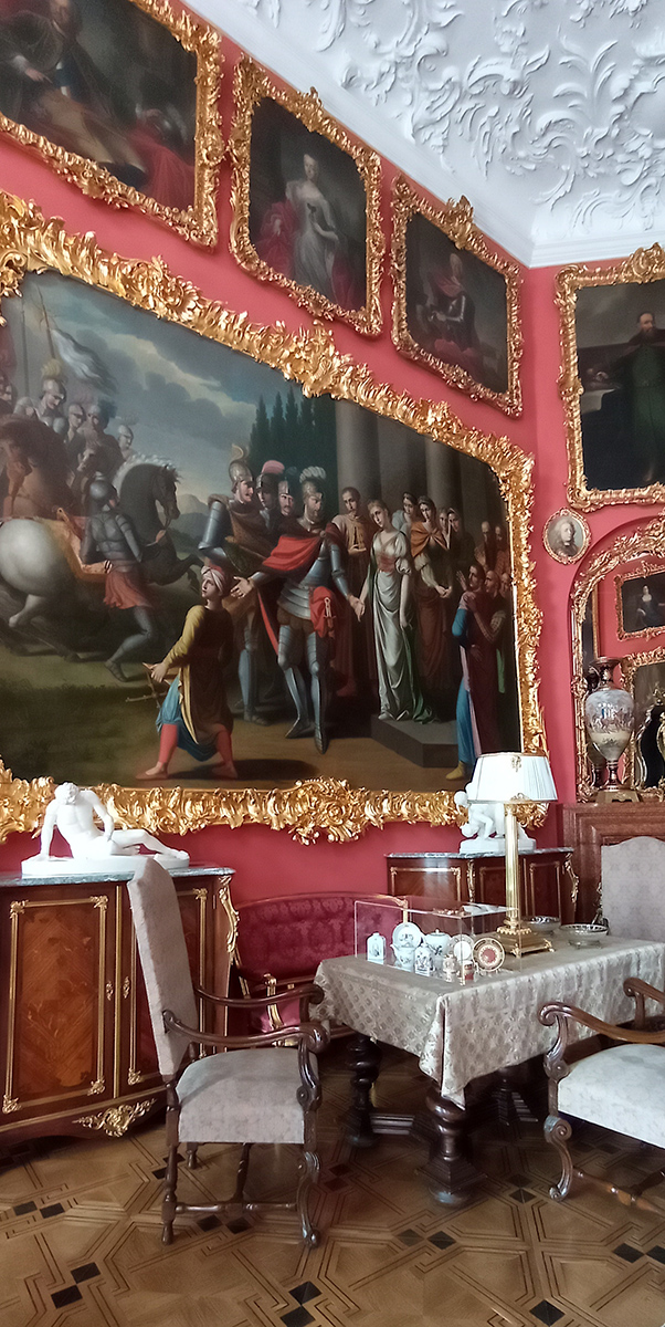 1. Salon Czerwony, ekspozycja do wystawy „Kredensie, otwórz się” Fotografia przedstawia fragment pomieszczenia, narożnik. Na pierwszym planie krzesło z siedziskiem i oparciem w jasnym kolorze oraz nóżkami i podłokietnikami w kolorze brązowym. Po prawej strony fragment takiego samego krzesła. Dalej stół z masywnymi nogami w kolorze ciemnego brązu nakryty obrusem w jasnym kolorze z ciemniejszym ornamentem. Na stole zestaw naczyń porcelanowych w przeźroczystym pudełku, za nimi lampa z białym abażurem i korpusem w kolorze złotym. Za stołem, pod ścianą kanapa z obiciem w kolorze ciemnoczerwonym. Po obu stronach kanapy szafki w kolorze brązowym ze zdobieniami w kolorze złotym. Na nich rzeźby w białym kolorze. Na ścianach obrazy w bogato zdobionych ramach w kolorze złotym. Największy z obrazów bezpośrednio nad kanapą. Podłoga drewniana w kolorze brązowym, z czarnymi wstawkami, z ornamentem geometrycznym.