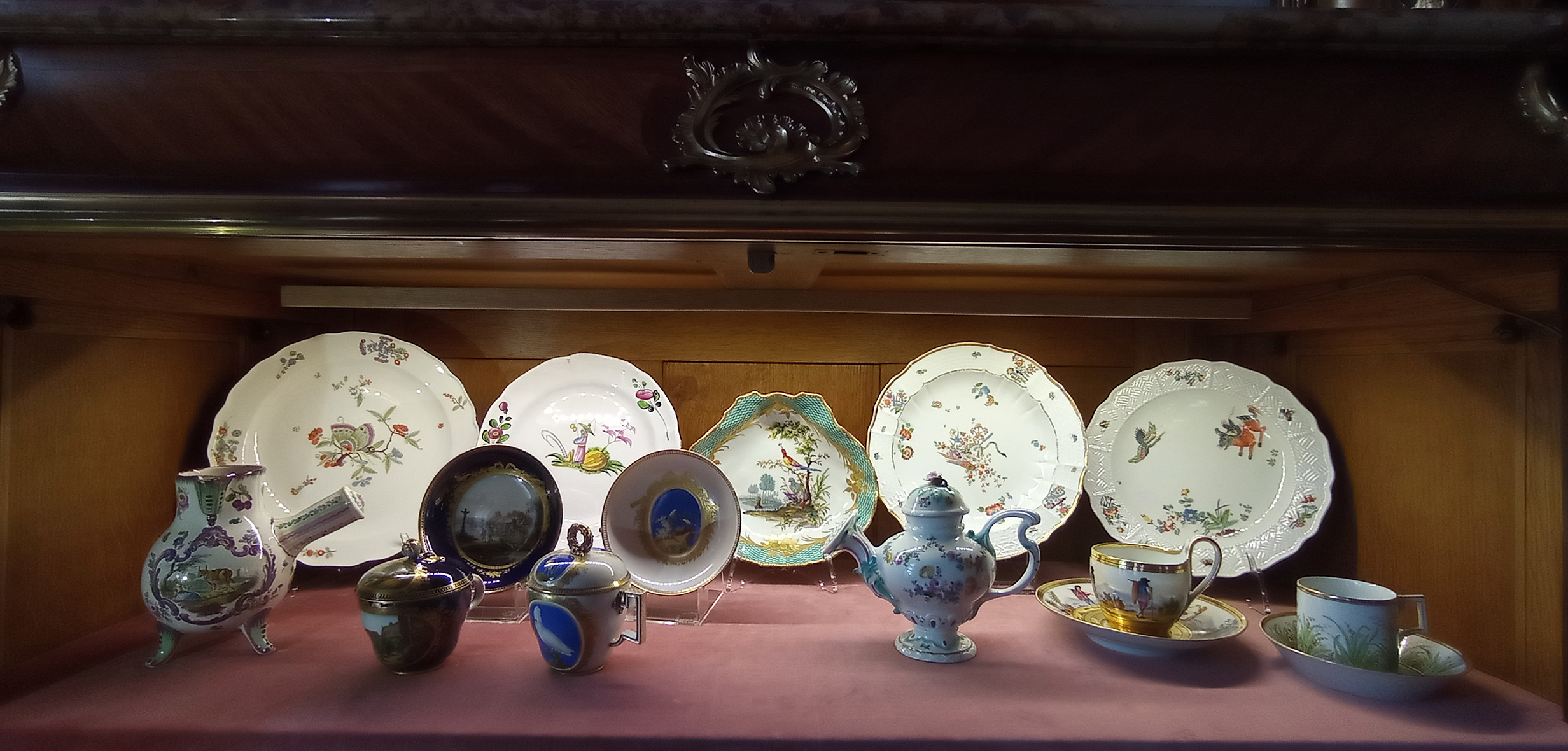 1. Górna półka witryny w Salonie Białym, ekspozycja do wystawy „Kredensie, otwórz się” Fotografia przedstawia wnętrze witryny z porcelanowymi naczyniami. Z tyłu pięć talerzy różnego rozmiaru i z różną dekoracją. Z przodu, od lewej. Naczynie o pękatym kształcie na trzech nóżkach, z dzióbkiem i uchwytem. W kolorze białym ze sceną figuralną i dekoracja roślinną. Dominuje kolor fioletowy. Obok zestaw filiżanka z przykrywką i spodkiem, spodek na podstawce. Filiżanka na niskiej podstawie, owoidalna, w połowie wysokości lekko wybrzuszona; z uszkiem; z przykrywką. Podstawa filiżanki złocona, korpus ciemnogranatowy, z wyodrębnionym owalnym polem, w nim scena: pejzaż z budynkami i myśliwym. Pole zamknięte złoconą taśmą z różami Krawędź podkreślona złoceniem. Uszko w formie przeplatających się pałączków, o złoconych grzbiecikach, w miejscach osadzenia przechodzących w stylizowane liście akantu cieniowane złotem. Przykrywka lekko wyniesiona, malowana ciemnogranatowo, z owalną kwaterą, w której pejzaż z zatoką, roślinnością, budowlą i skałami w tle. Na skale, na pierwszym planie, stojący chłopczyk. Podobna dekoracja na spodku. Obok kolejny zestaw, filiżanka i spodek na podstawce. Filiżanka owoidalna, lekko wybrzuszona, z uchem załamanym pod kątem prostym. Przykrywka wysklepiona, z uchwytem w formie wieńca laurowego. Na korpusie filiżanki, w polu, na błękitnym tle, po jednej stronie biała papuga z koszykiem w dziobie, siedząca na gałęzi. Na przykrywce miniatura z żółtym ptaszkiem. Wokół brzegu złota plecionka z zachodzących na siebie kółek. Podobny motyw na spodku. Obok naczynie do pudrowania ciast na okrągłej stopie, z krótkim trzonem; z korpusem w kształcie odwróconej gruszki, przykryte kopulastą, zakręconą przykrywką; z wygiętym uchem i dziobkiem zakończonym okrągłym, płaskim sitkiem. Dekoracja roślinna. Obok zestaw filiżanka stojąca na spodku. Dekoracja w kolorze złotym, ze scenami rodzajowymi. Na filiżance mężczyzna wsparty na kiju, w tle pejzaż górski, kamienie i krzewy. Skrajnie po prawej zestaw filiżanka stojąca na spodku. Na nich złocenia, dekoracja roślinna w kolorach żółtym, czerwonym, niebieskim i różnych odcieniach zieleni.
