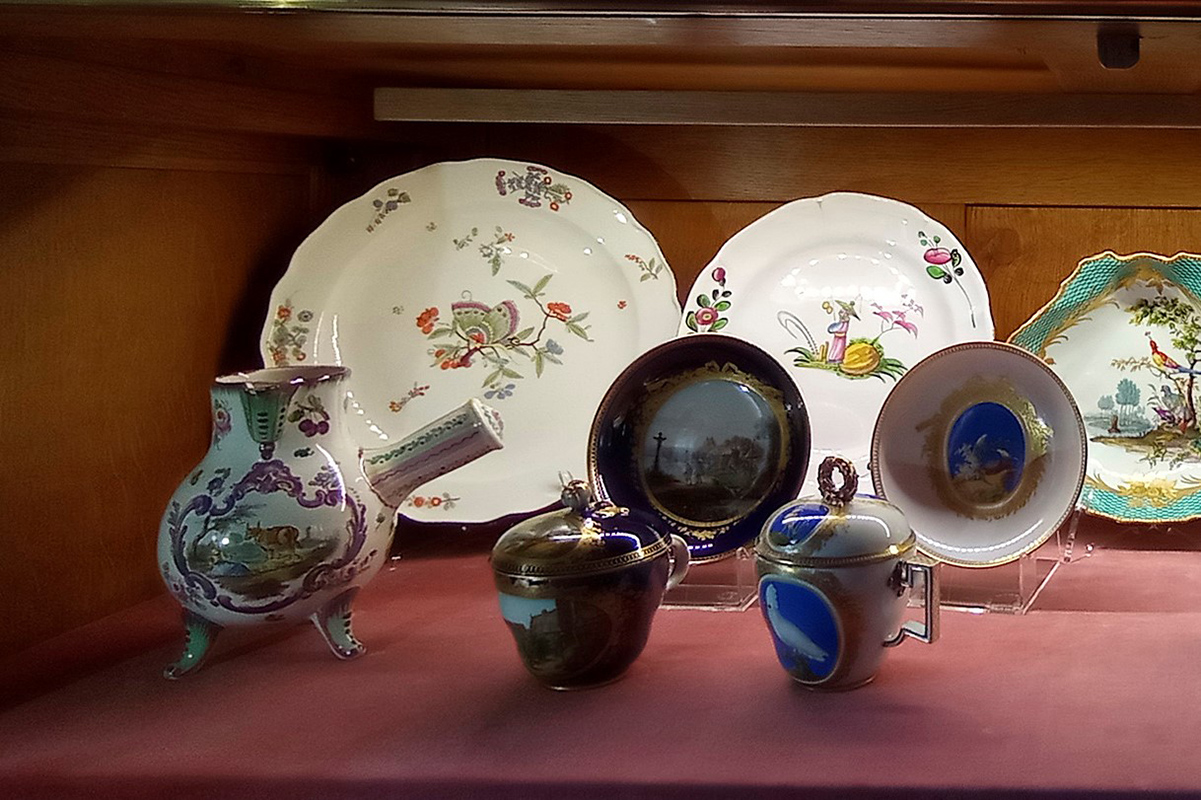 Naczynia do picia czekolady: dzbanuszek i dwie filiżanki ze spodkami. Witryna w Salonie Białym / Chocolate drinking utensils: a jug and two cups and saucers. Showcase in the White Salon Fotografia przedstawia wnętrze witryny z porcelanowymi naczyniami. Z tyłu trzy talerze różnego rozmiaru i z różną dekoracją. Z przodu, od lewej. Naczynie o pękatym kształcie na trzech nóżkach, z dzióbkiem i uchwytem. W kolorze białym ze sceną figuralną i dekoracja roślinną. Dominuje kolor fioletowy. Obok zestaw filiżanka z przykrywką i spodkiem, spodek na podstawce. Filiżanka na niskiej podstawie, owoidalna, w połowie wysokości lekko wybrzuszona; z uszkiem; z przykrywką. Podstawa filiżanki złocona, korpus ciemnogranatowy, z wyodrębnionym owalnym polem, w nim scena: pejzaż z budynkami i myśliwym. Przykrywka lekko wyniesiona, malowana ciemnogranatowo. Podobna dekoracja na spodku. Obok kolejny zestaw, filiżanka i spodek na podstawce. Filiżanka owoidalna, lekko wybrzuszona, z uchem załamanym pod kątem prostym. Przykrywka wysklepiona, z uchwytem w formie wieńca laurowego. Na korpusie filiżanki, w polu, na błękitnym tle biała papuga z koszykiem w dziobie, siedząca na gałęzi. Na przykrywce miniatura z żółtym ptaszkiem. Wokół brzegu złota plecionka z zachodzących na siebie kółek. Podobny motyw na spodku.