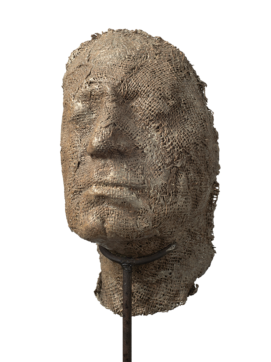 Magdalena Abakanowicz, Portret anonimowy Magdalena Abakanowicz
1930 Falenty – 2017 Warszawa
Portret anonimowy
2000
Żywica metal (stelaż)
103 x 22 x 23
Starak Collection
Praca ukazuje silnie uproszczoną, niemal ascetyczną głowę – rzeźbę wykonaną z żywicy o powierzchni przypominającej spękaną, wyschniętą ziemię. Materiał ten nadaje formie porowatą, kruchą strukturę, która zdaje się być świadectwem upływu czasu, przemijania i organicznej ulotności.
Głowa jest zamocowana na długim, cienkim metalowym pręcie, który osadzony jest w minimalistycznej, kwadratowej podstawie. Ten kontrast między miękką, niemal organiczną materią a zimnym, surowym metalem podkreśla napięcie między cielesnością a abstrakcją, między ludzkim doświadczeniem a mechaniczną formą nośną.
Twarz – pozbawiona większości szyi, jakby wyjęta z ciała i zawieszona w przestrzeni – emanuje głęboką emocją. Zamknięte lub przymknięte oczy otoczone są wyraźnymi bruzdami, które mówią o cierpieniu, melancholii lub intensywnej zadumie. Usta lekko opuszczone podkreślają powagę, a może nawet ból, tworząc wyraz pełen dramatyzmu i skupienia.
Rzeźba zdaje się nie tylko przedstawiać postać, ale raczej wprowadzać widza w przestrzeń emocjonalną, gdzie surowość materiału i minimalizm formy stają się nośnikiem uniwersalnej ludzkiej kondycji.