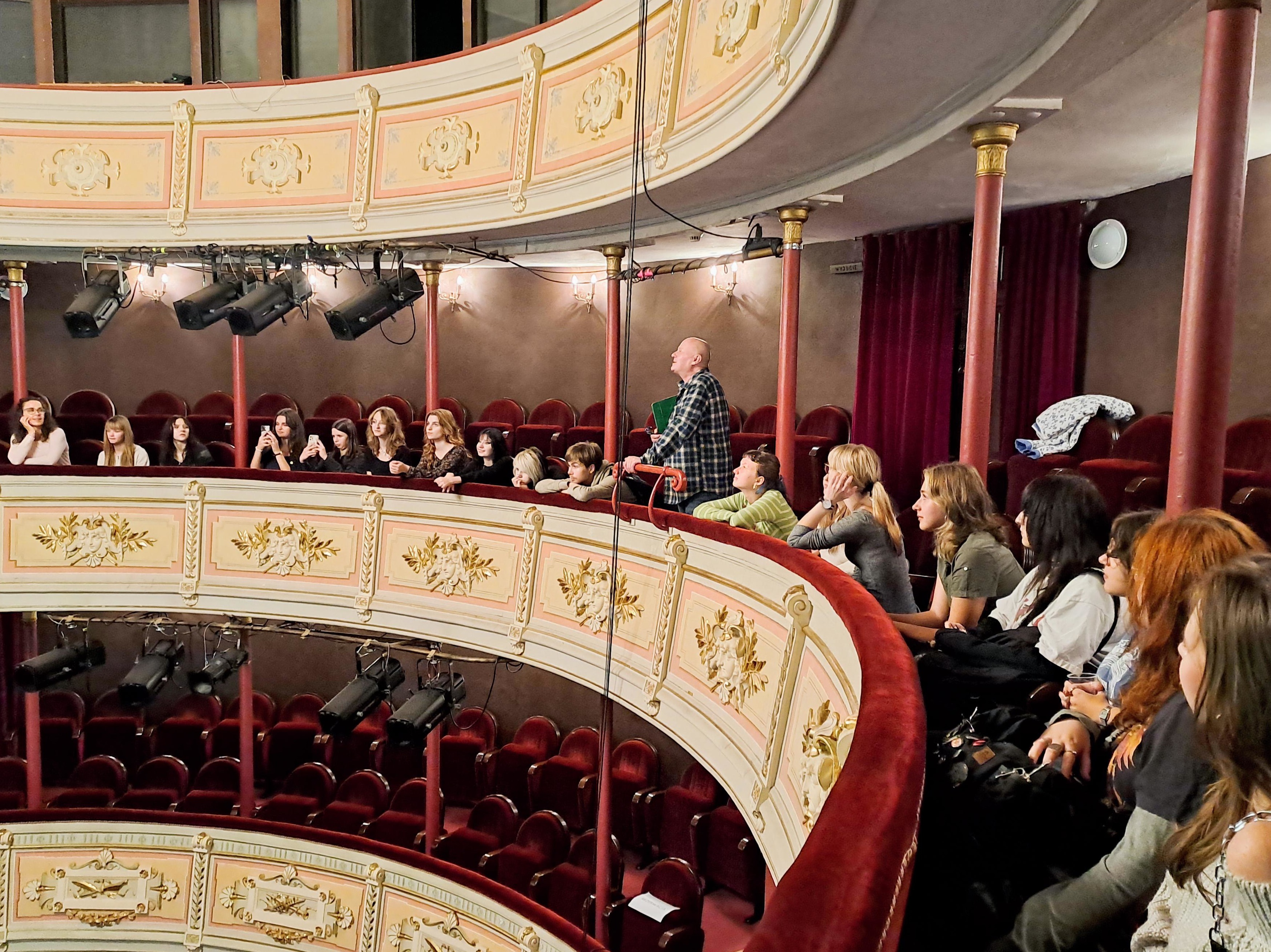 Na zdjęciu widać grupę młodzieży wraz z dorosłym opiekunem, pozujących wspólnie na tle teatralnej scenografii.
Osoby siedzą i stoją na schodach ustawionych przed sceną. Grupa liczy około dwudziestu osób – większość to nastolatki, głównie dziewczęta, a wśród nich jeden starszy mężczyzna w koszuli w kratę. Wszyscy patrzą w stronę obiektywu, część uśmiecha się, inni mają poważniejsze miny.
W tle widoczna jest teatralna scenografia – dwa duże, malowane elementy przypominające fragmenty antycznej architektury z łukami. Widać na nich pejzaże: niebo, wodę, drzewa i budynki. Nad sceną rozciągnięto jasną siatkę lub materiał przypominający baldachim.
Cała grupa sprawia wrażenie zadowolonej, jakby zdjęcie było pamiątką po wspólnym wydarzeniu – najpewniej wizycie w teatrze lub warsztatach scenicznych.