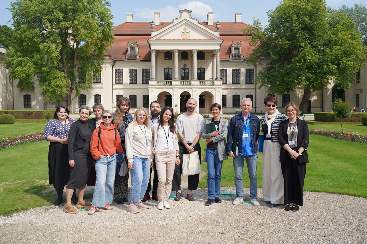 Grupa trzynastu osób stoi przed pałacem w Kozłówce na tle zielonego ogrodu. Uczestnicy uśmiechają się do obiektywu. W tle widać jasną fasadę pałacu z kolumnowym tarasem i czerwonym dachem. Zdjęcie wykonano w słoneczny, letni dzień.