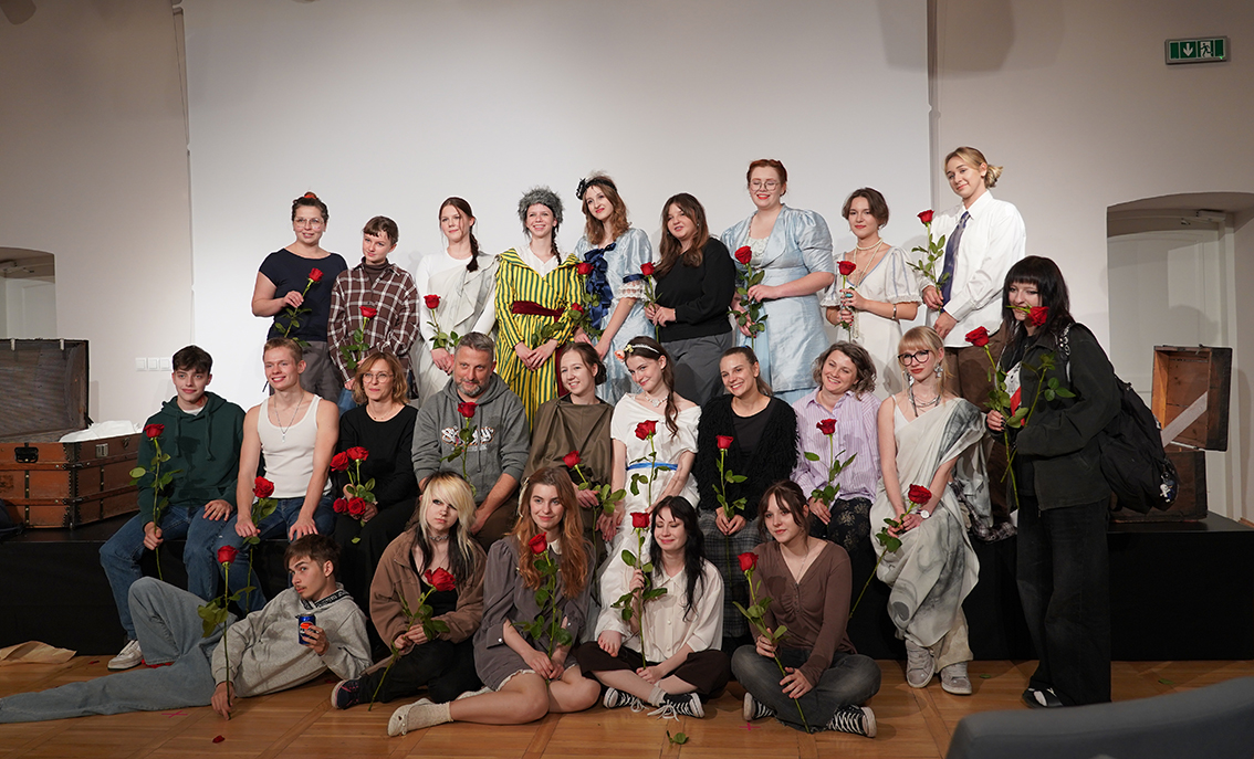 Na zdjęciu widzimy dużą, około dwudziestoosobową grupę młodych osób i dorosłych, pozujących wspólnie na scenie po zakończonym występie. Większość z nich siedzi lub stoi w kilku rzędach, uśmiechając się do obiektywu. W rękach trzymają pojedyncze, długie czerwone róże – symbol podziękowania i zakończenia projektu.

Część uczestników ubrana jest w codzienne stroje, inni mają na sobie kostiumy teatralne w stylu historycznym – długie suknie, eleganckie kamizelki, ozdobne dodatki. W tle widać jasną ścianę i sceniczne oświetlenie, po bokach stoją elementy scenografii – duże, drewniane kufry. Atmosfera zdjęcia jest radosna i pełna dumy po udanym przedstawieniu.