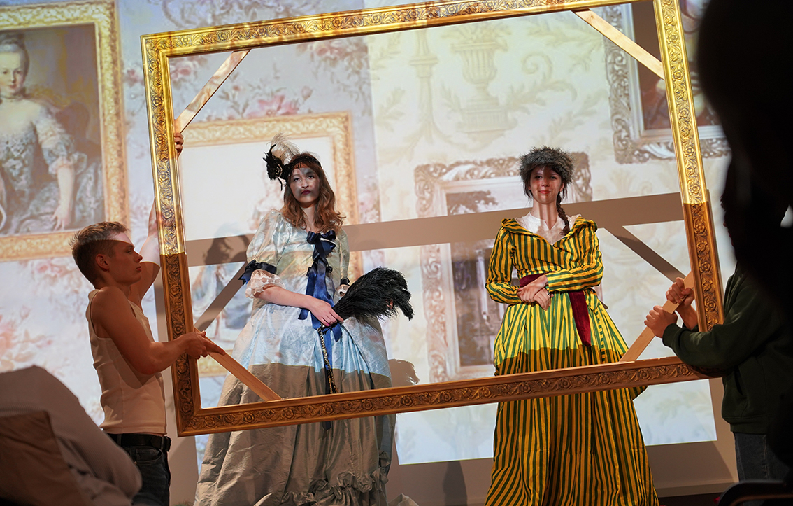 Na zdjęciu widać scenę przedstawienia inspirowanego dawnym malarstwem. Na pierwszym planie dwie młode osoby stoją wewnątrz dużej, złotej ramy obrazu, którą po bokach trzymają inni uczestnicy spektaklu.
Postać po lewej stronie ma na sobie błękitną, długą suknię z bufami i ciemnoniebieskimi wstążkami, na głowie czarną ozdobę z piórami. W dłoniach trzyma czarne pióro – wachlarz. Obok niej stoi osoba w żółto-zielonej sukni w pionowe pasy, z białym kołnierzem i czerwonym pasem w talii; na głowie ma futrzaną czapkę.
W tle wyświetlone są powiększone reprodukcje obrazów przedstawiających kobiety w strojach z XVIII wieku w złoconych ramach. Całość tworzy wrażenie, że aktorki stały się częścią ożywionego obrazu.
Scena jest barwna i teatralna – światło skupia się na postaciach w ramie, podkreślając efekt „żywego obrazu”.
