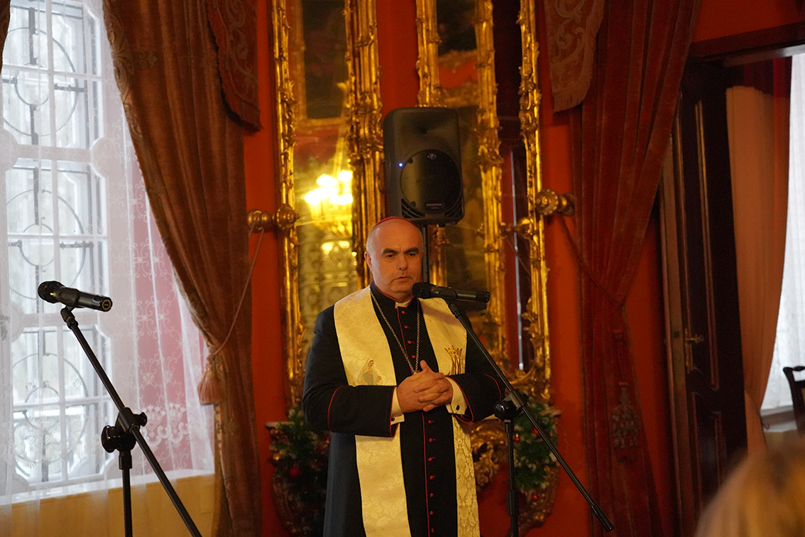 Zdjęcie przedstawia uroczyste wystąpienie w bogato zdobionym, pałacowym wnętrzu. Tłem są czerwone ściany, wysokie okna z białymi firanami oraz ciężkie, bordowe zasłony. Za centralną postacią widoczne są duże, pionowe lustra w złoconych ramach, odbijające ciepłe światło lamp i podkreślające reprezentacyjny charakter sali.
W centrum kadru stoi duchowny w czarnej sutannie z czerwonymi obszyciami. Na ramionach ma jasną, kremowo-złotą stułę z delikatnymi haftami. Jego dłonie są złożone na wysokości klatki piersiowej. Twarz ma skupioną i poważną, a wzrok skierowany przed siebie.
Przed nim znajduje się mikrofon na statywie, drugi mikrofon ustawiony jest nieco z boku po lewej stronie kadru. Za postacią widoczny jest czarny głośnik. U dołu, przy ścianie, stoją zielone dekoracje roślinne.
Całość sceny sprawia wrażenie podniosłej chwili — przemówienia lub modlitwy — wygłaszanej podczas oficjalnego wydarzenia w eleganckiej, historycznej przestrzeni.