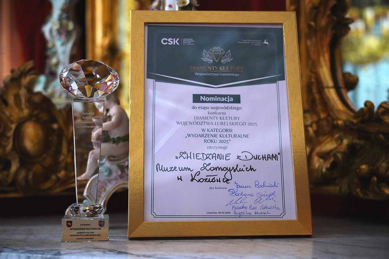 Na fotografii widoczne są dwa elementy ustawione obok siebie na stole: szklana statuetka w kształcie diamentu oraz dyplom w złotej ramie.
Po lewej stronie stoi wysoka, przezroczysta statuetka. Na jej szczycie znajduje się duży, oszlifowany kryształ przypominający diament. Pod nim znajduje się smukła, szklana podstawa z wygrawerowanym napisem. Na dolnym elemencie podstawy umieszczona jest niewielka tabliczka z informacją o kategorii nagrody – „Wydarzenie kulturalne”.
Po prawej stronie znajduje się dyplom w szerokiej, złotej ramie. W górnej części dokumentu widoczne jest logo CSK oraz napis „Diamenty Kultury Województwa Lubelskiego”. W centralnej części dyplomu znajduje się informacja o nominacji do etapu wojewódzkiego w konkursie w kategorii „Wydarzenie kulturalne roku 2025”. Niżej odręcznym pismem wpisano tytuł wydarzenia „Zwiedzanie z duchami” oraz nazwę instytucji – Muzeum Zamoyskich w Kozłówce. Na dole widoczne są podpisy członków jury.
Tło fotografii jest miękko rozmyte. Widać fragmenty bogato zdobionego wnętrza pałacowego – złocone ornamenty i dekoracyjne elementy wyposażenia, które podkreślają reprezentacyjny charakter miejsca. Całość utrzymana jest w ciepłych, złotych i czerwonych tonach, co nadaje zdjęciu elegancki, uroczysty nastrój.